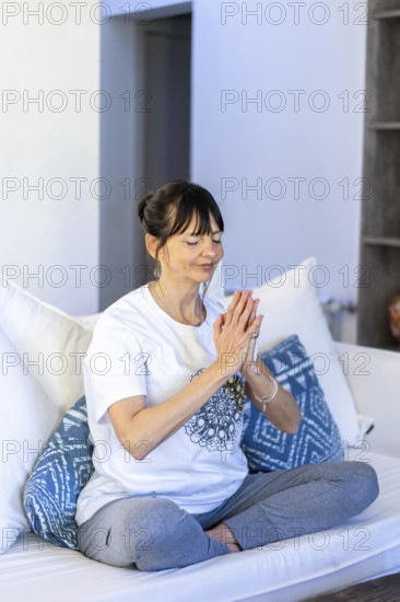 Mid-adult woman meditating on her sofa at home, with natural daylight and palms pressed together at heart center