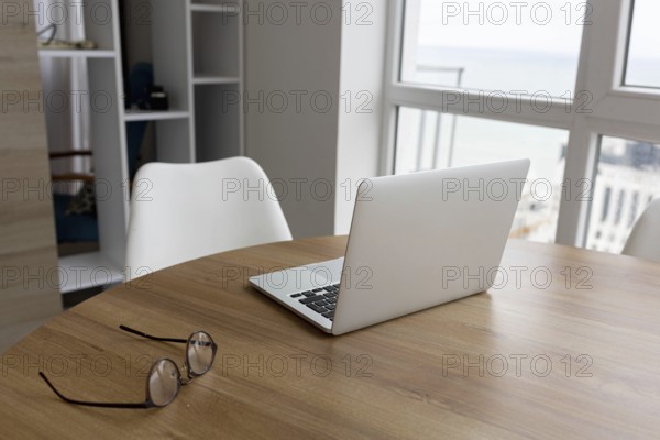 A wooden table with a laptop and glasses next to a white chair in a bright room