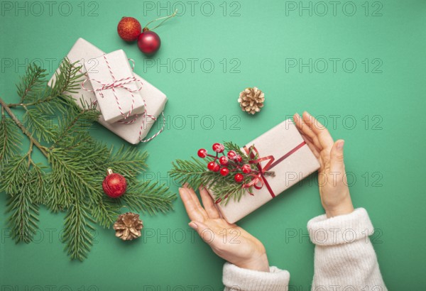 Christmas or New Year celebration green paper festive background with female hands holding wrapped gift box, decoration fir tree, cones, berries, sparkly red balls. Giving presents concept, food photography, food photography