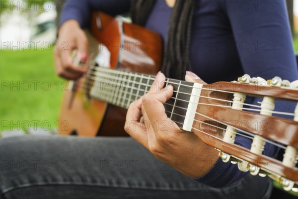 Woman with Brown Skin and Long Black Hair with Dreadlocks Sitting and Playing Guitar in Green Park. Medium Shot