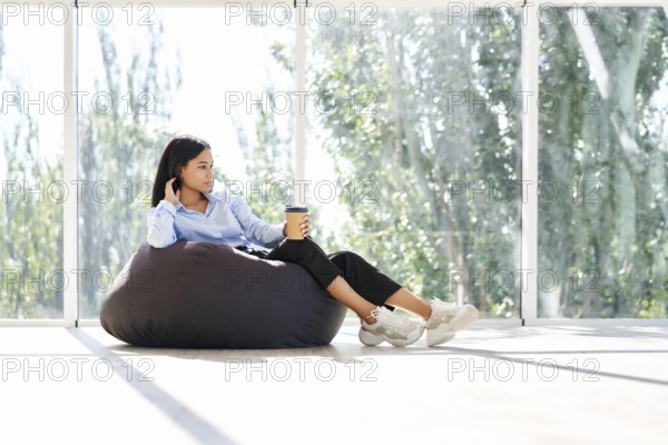 Pretty african american business woman relaxing on bean bag chair with cup of coffee on panoramic windows background in modern creative office with copy space. Break time concept, business success