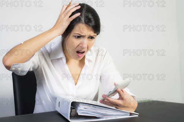 Businesswoman sitting at her desk with a large office binder looking at her calculator in horror or dismay with her mouth open in shock