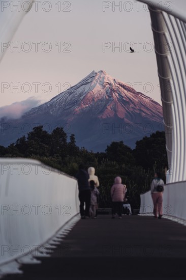 People on a bridge admiring a snow-capped mountain at sunset with a bird in the sky, Mount Taranaki, New Zealand, Oceania