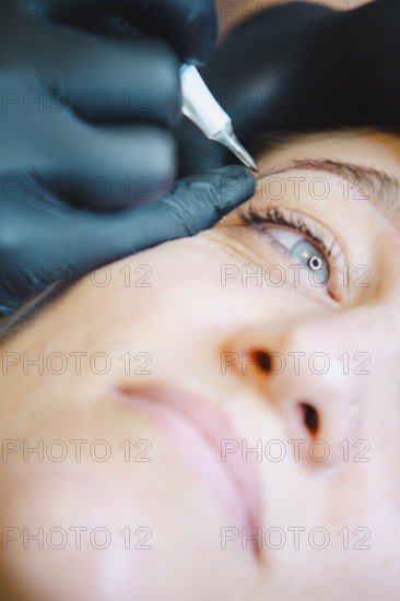 Beautician applies permanent makeup to a woman's eyebrow in a busy salon, showcasing precision and skill