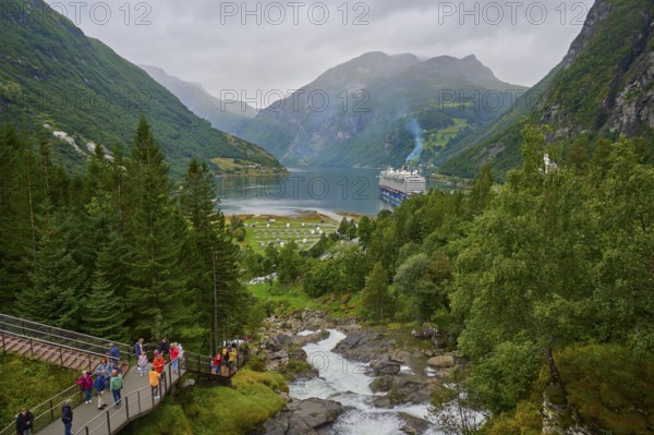 An impressive landscape with a raging waterfall, surrounded by green mountains and forests, with a cruise ship, Mein Schiff 6, on a fjord hiking trail with walkway and tourists, Geiranger, Geiranger Fjord, Stranda, Romsdal, Norway, Europe