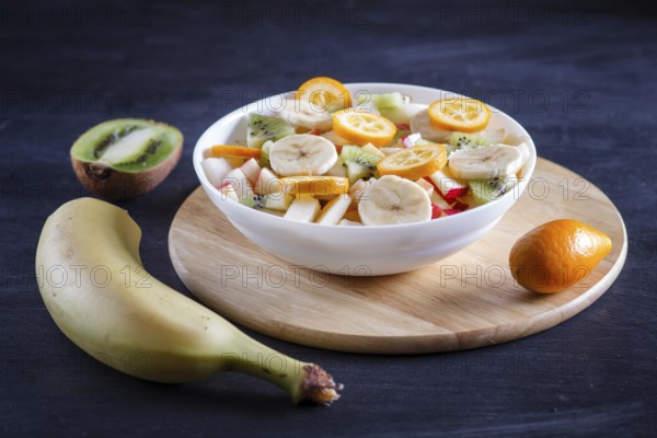 Vegetarian salad of bananas, apples, pears, kumquats and kiwi on black woodem background, close up, selective focus