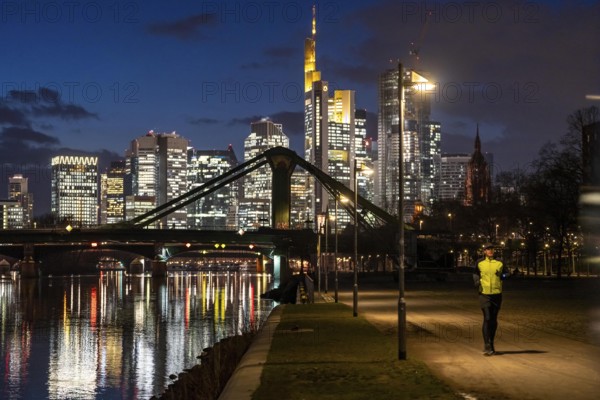 Skyline of the city centre of Frankfurt am Main, joggers on the pavement, promenade along the river Main, dusk, Hesse, Germany raft bridge in the background