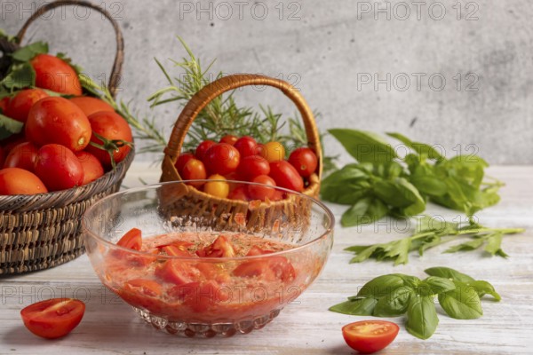 Glass bowl with prepared tomato sauce and sliced tomatoes, surrounded by fresh tomatoes in baskets, basil and rosemary