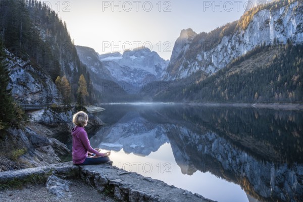 A woman doing yoga by the lake. Lotus position (Padmasana) . Vorderer Gosausee in autumn with a view of the Dachstein mountains. The Gosaukamm on the right. Blue sky, good weather. Reflection. Morning atmosphere. Vorderer Gosausee, Gosau, Gosau valley, Salzkammergut, Upper Austria, Austria, Europe