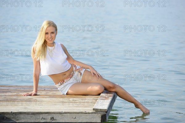 Blonde woman in a white top and shorts lying down relaxed on the dock by the lake, Austria, Europe