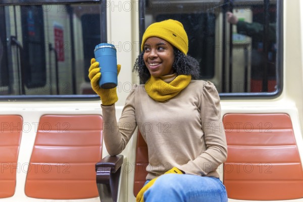 Smiling young woman wearing yellow winter accessories holding reusable mug while commuting on public transport