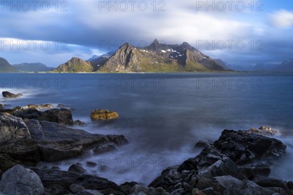 Landscape with sea and mountains on the Lofoten Islands, view across the fjord to the small town of Flakstad and the mountain Flakstadtinden as well as other mountains. Rocks in the foreground. At night at the time of the midnight sun in good weather, some clouds in the sky. Long exposure, blurred clouds. Early summer. Flakstadoya, Lofoten, Norway, Europe