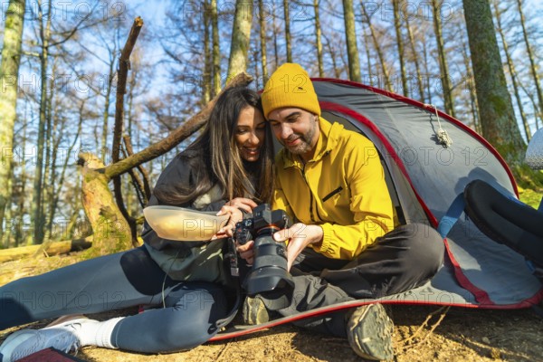 Couple of hikers reviewing photos on professional camera while having a meal in front of their tent during camping trip in forest