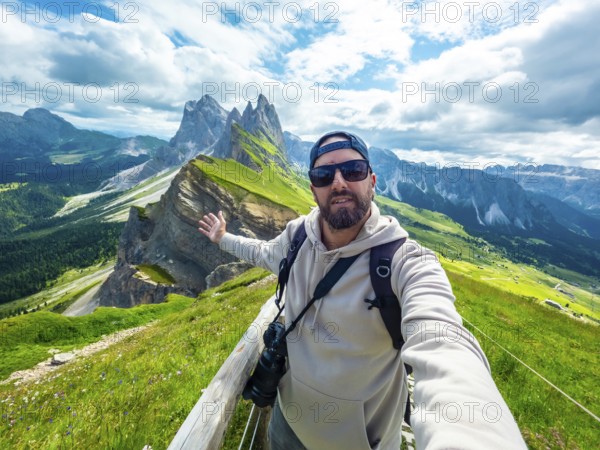 Photographer taking a selfie with outstretched arm, capturing the breathtaking seceda peaks and lush valleys of the dolomites during a sunny summer day