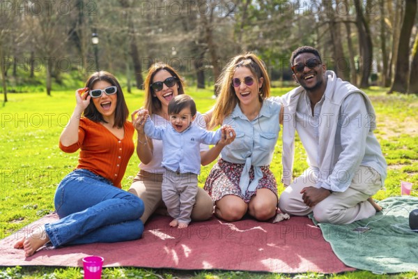 Cheerful multi ethnic friends enjoying a fun picnic in the park, laughing and bonding together with a baby on a sunny day