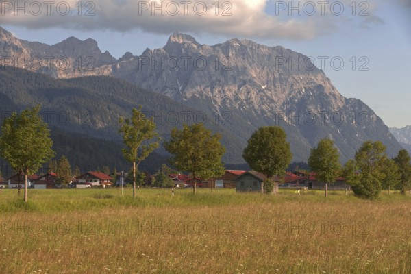 View of a village in front of majestic mountains, surrounded by green meadows and trees under a cloudy sky, Wallgau, Bavaria, Germany, Europe