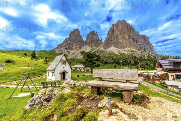 Picturesque landscape featuring a wooden bench, a small chapel, and the majestic peaks of the sella group in the italian dolomites during summer