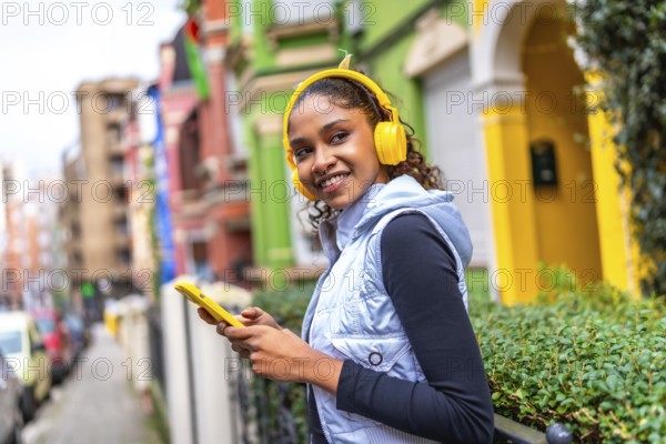 Confident african american woman using phone and listening to music walking next a multicolored houses in the city
