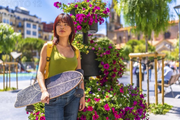 Portrait with copy space of a young chinese woman carrying skateboard on a city square full of purple flowers