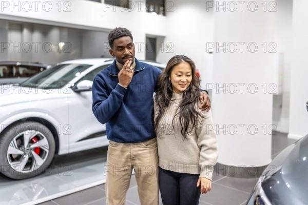 Happy multiethnic couple choosing their new electric car in a dealership showroom, sustainable mobility and green transportation concept