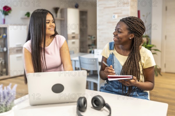 Two smiling teenage girls collaborating on school project, using laptop and taking notes, enjoying studying together at home
