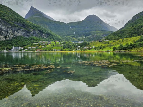 Mountainous landscape with a calm fjord reflecting the surrounding mountains and village, surrounded by green nature and under a cloudy sky, Geiranger, Geiranger Fjord, Stranda, Romsdal, Norway, Europe