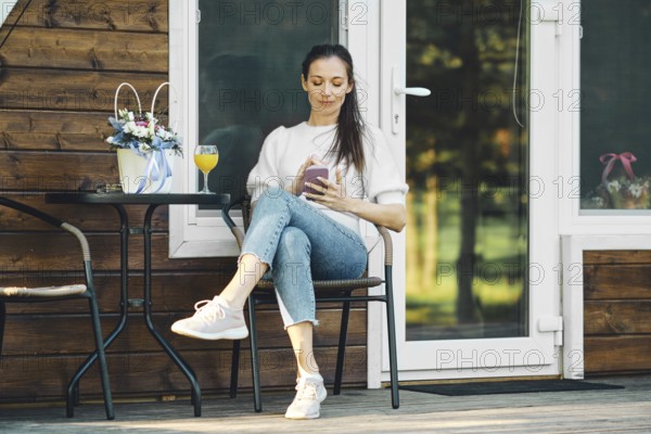 Relaxed woman sitting on terrace of wooden a-frame cabin with smartphone