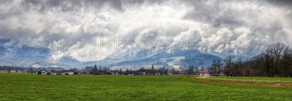 Green meadow and small village in the foreground, dramatic clouds, mountains in the background, Bad Feilnbach