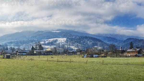 A village in front of snow-covered mountains under a cloudy sky on a green meadow, Bad Feilnbach, winter