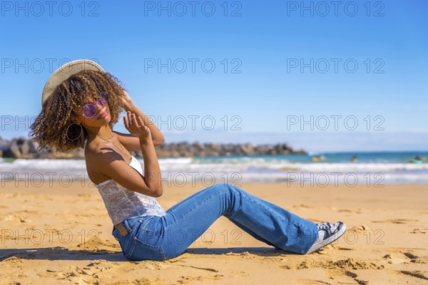 Beauty latin woman sitting on the sand of a beach playing with sun hat and smiling at camera