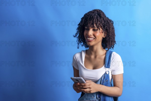 Young woman smiling while using a smartphone against a vibrant blue background, showcasing a modern lifestyle and connection through technology