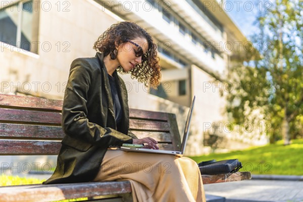 Profile of a beauty young arab businesswoman in casual clothes and sunglasses working with laptop sitting on park bench