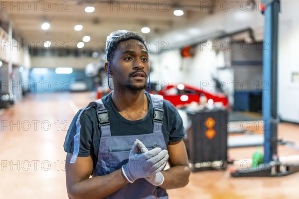 Confident young mechanic wearing gloves and protective glasses stands in a professional auto repair shop, ready to work