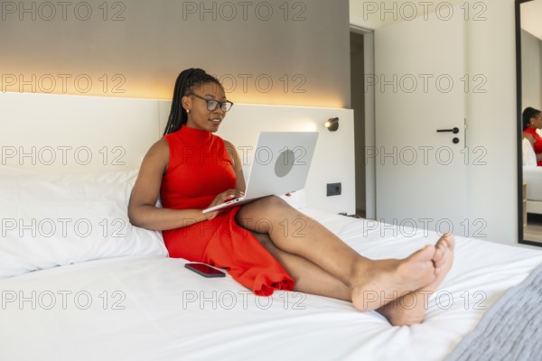 Businesswoman wearing a red dress using a laptop while sitting comfortably on a hotel bed during a business trip