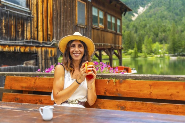 Woman with straw hat enjoying a hot drink in a bar with mountain lake view in the dolomites