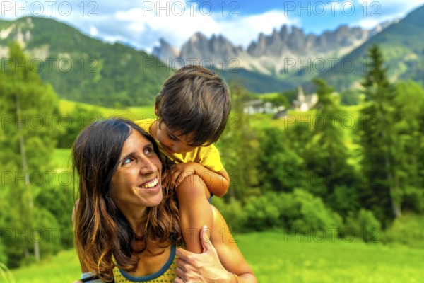 Mother carrying her son on her shoulders, enjoying a sunny day in the italian dolomites with the church of st. Magdalena nearby