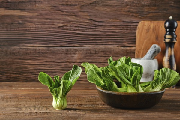 A bunch of vibrant bok choy sits in a bowl alongside a single piece on a wooden table, complemented by a mortar and pestle and a pepper mill, suggesting a cozy cooking atmosphere.