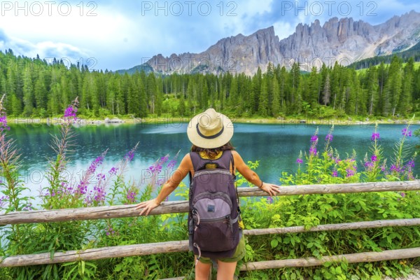 Female tourist with backpack admiring the stunning turquoise waters of lake carezza and the latemar mountain range in the italian dolomites during summer