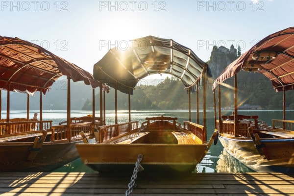 Wooden tourist boats moored at dock on lake bled in slovenia with bled island and iconic church in background and sun shining through canopy