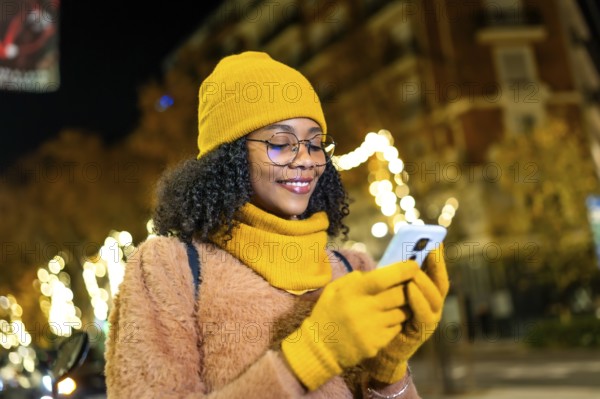 Smiling young woman wearing yellow winter clothes using smartphone in a city street at night with christmas lights