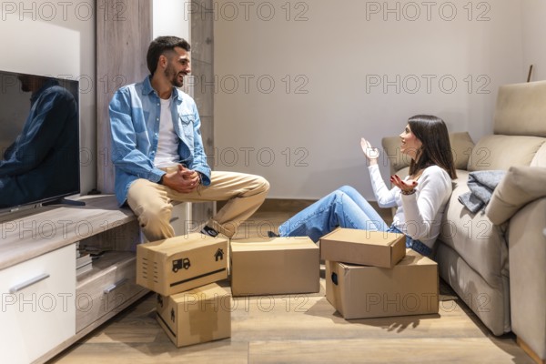 Young couple talking and smiling while unpacking cardboard boxes in new home, planning the arrangement of furniture