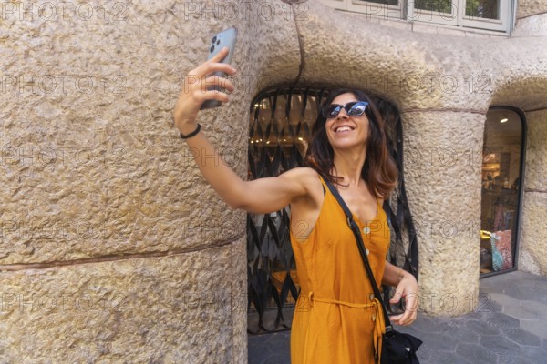Smiling woman taking selfie with smartphone in front of casa mila in barcelona, enjoying summer tourism in catalonia