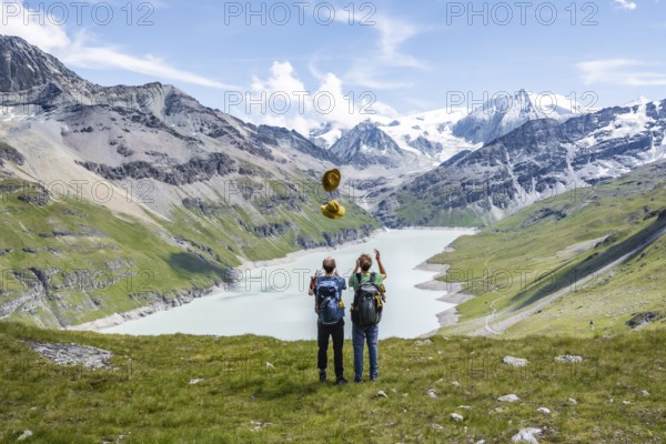 Two friends throwing yellow hats in the air, mountaineers in front of a blue mountain lake, Lac des Dix reservoir, behind mountain peak Mont Blanc de Cheilon with glacier, Hérménence, Valais Alps, Valais, Switzerland, Europe
