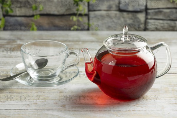 A teapot with red tea next to an empty glass cup with spoon on a table in front of a stone background