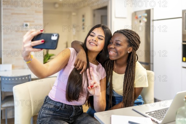 Two cheerful teenage girls enjoying a fun moment while taking a selfie with a smartphone, studying together at home