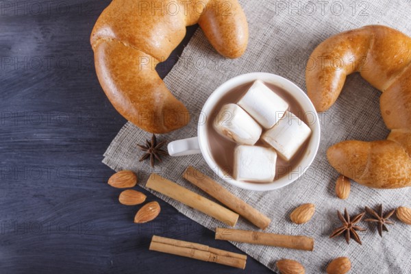 A cup of hot chocolate with marshmallow, croissants and spices on linen napkin and black wooden background. top view, copy space