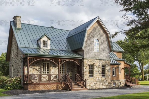 Old 1826 Canadiana cottage style fieldstone house facade with vertical wood plank cladding, cedar shingles and green standing seam sheet metal roof in late summer, Quebec, Canada, North America