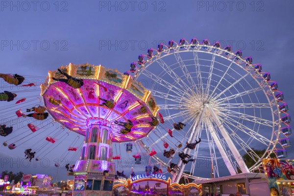 A lively funfair at dusk, illuminated by a large Ferris wheel and a colourful chain carousel, Cannstadter Volksfest, Stuttgart-Bad Cannstadt, Baden-Württemberg