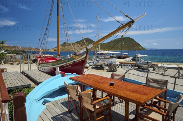 A harbour scene with wooden boats and a table with chairs and a blue boat in the foreground in sunny weather, Patmos Marine, boatyard, Diakofti, Patmos, Dodecanese, Greek Islands, Greece, Europe