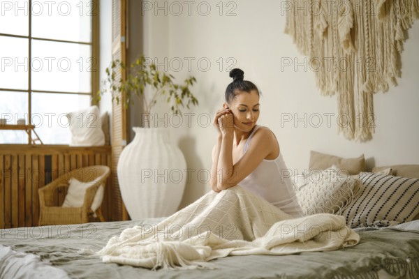 Peaceful woman sits on bed with her legs covered by cozy plaid. She is looking to the side and thinking about her fantasies